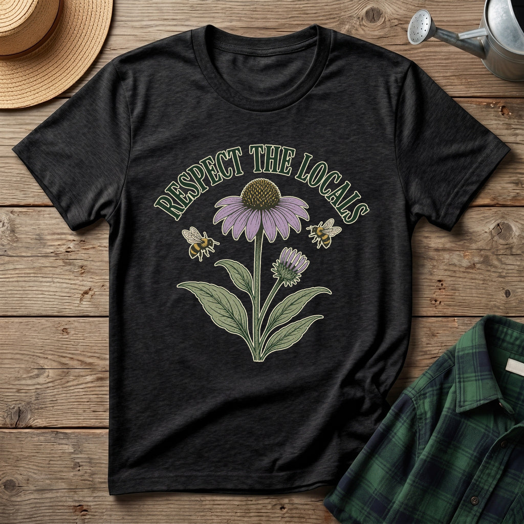 A black graphic t-shirt laid flat on a rustic wooden surface, featuring a vintage-style illustration of a purple coneflower, green leaves, two bees, and the text 'RESPECT THE LOCALS'. A straw hat, a metal watering can, and a green plaid shirt are styled around it.