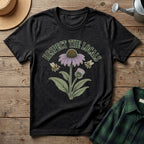 A black graphic t-shirt laid flat on a rustic wooden surface, featuring a vintage-style illustration of a purple coneflower, green leaves, two bees, and the text 'RESPECT THE LOCALS'. A straw hat, a metal watering can, and a green plaid shirt are styled around it.