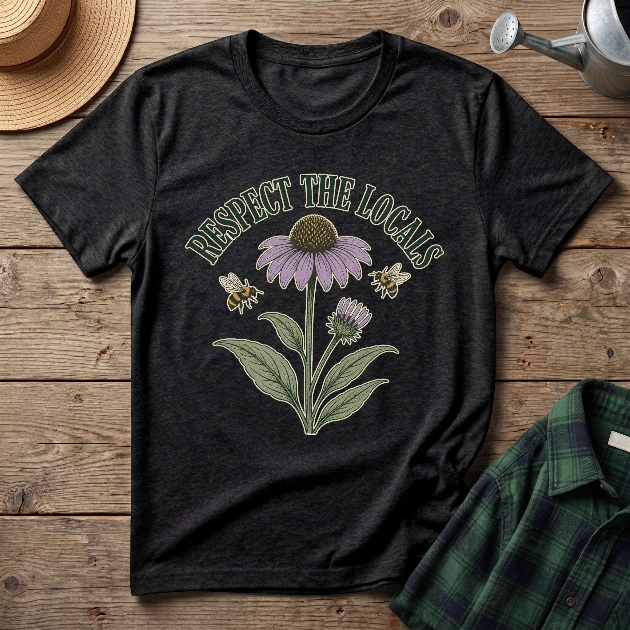 A black graphic t-shirt laid flat on a rustic wooden surface, featuring a vintage-style illustration of a purple coneflower, green leaves, two bees, and the text 'RESPECT THE LOCALS'. A straw hat, a metal watering can, and a green plaid shirt are styled around it.