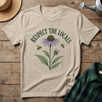 A beige graphic t-shirt laid flat on a rustic wooden surface, featuring a purple coneflower, green leaves, two bees, and the text 'RESPECT THE LOCALS' in dark green. A straw hat, metal watering can, and green plaid shirt are styled around it.