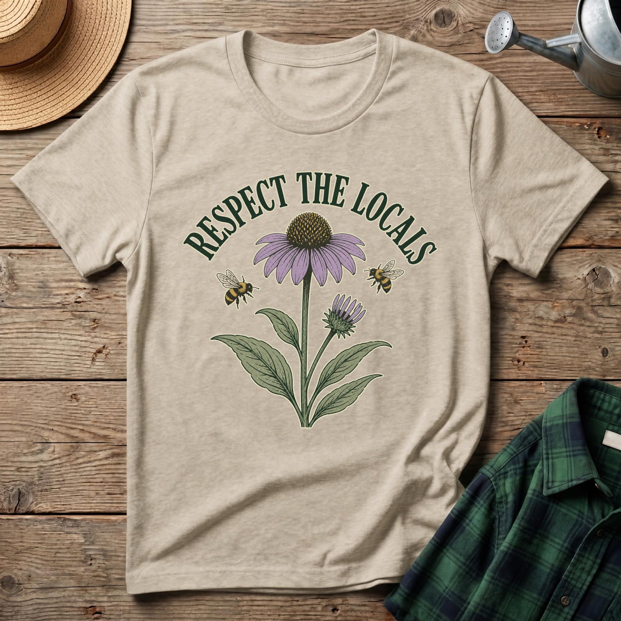 A beige graphic t-shirt laid flat on a rustic wooden surface, featuring a purple coneflower, green leaves, two bees, and the text 'RESPECT THE LOCALS' in dark green. A straw hat, metal watering can, and green plaid shirt are styled around it.